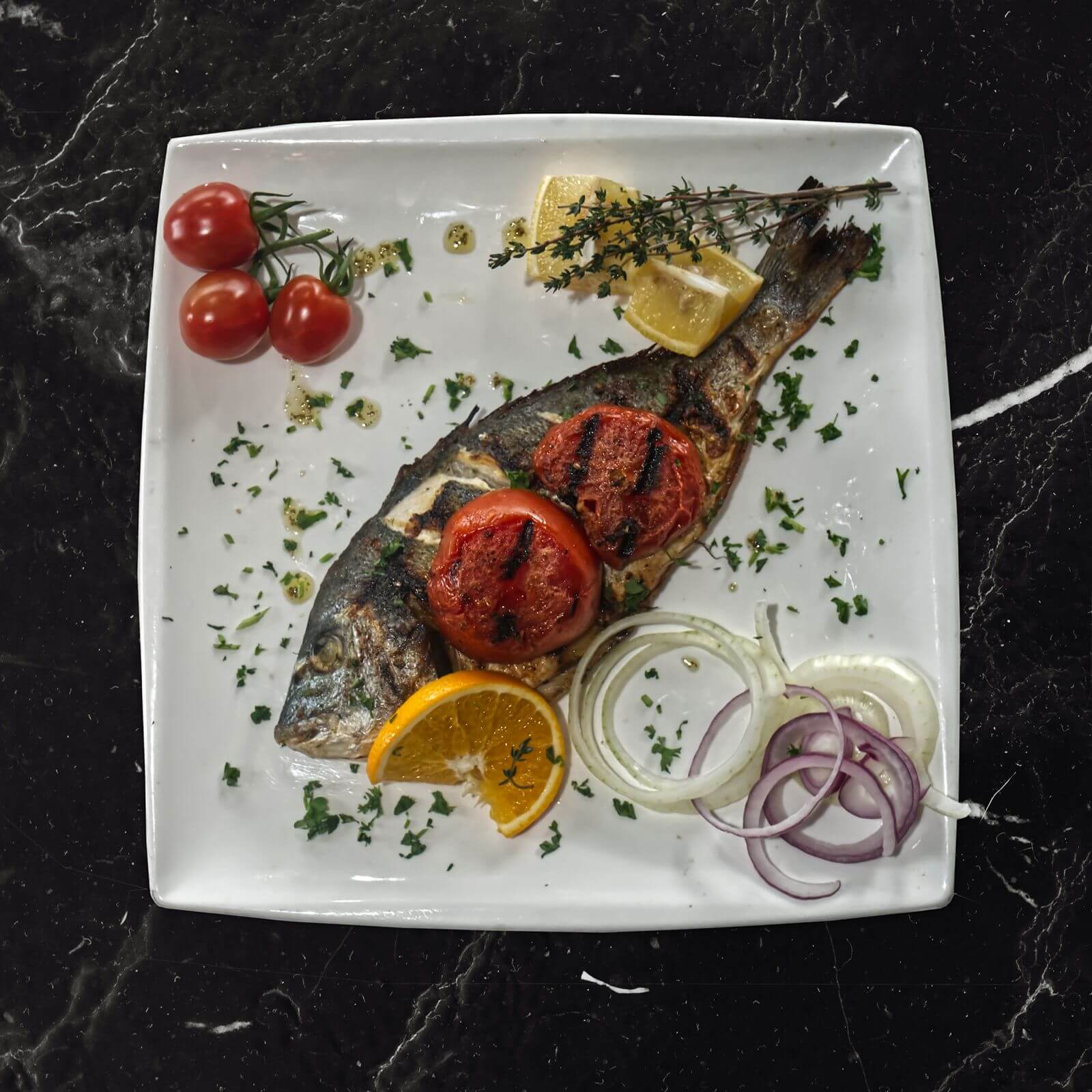 Gegrilde dorade op een wit bord met gegrilde tomaat, citroen, sinaasappel, tijm en uienringen.