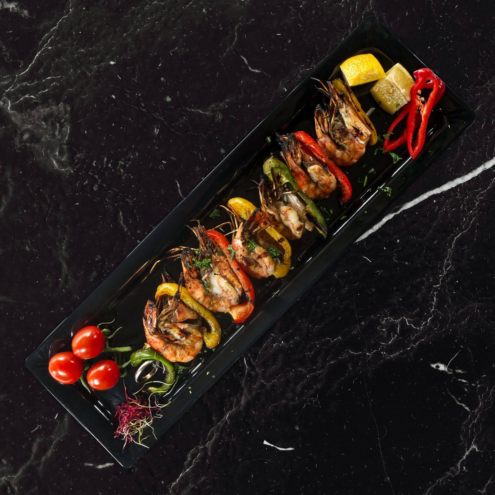 Spiesen met gegrilde grote gamba’s en paprika op een zwart langwerpig bord met citroen en tomaatjes.