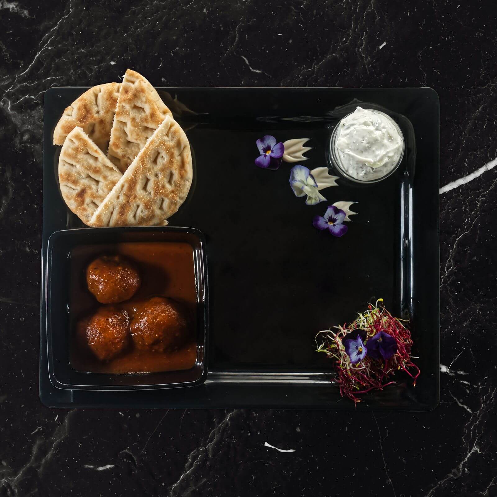 Keftedakia: gehaktballetjes in tomatensaus met pitabrood, tzatziki en eetbare viooltjes op een zwarte plaat.