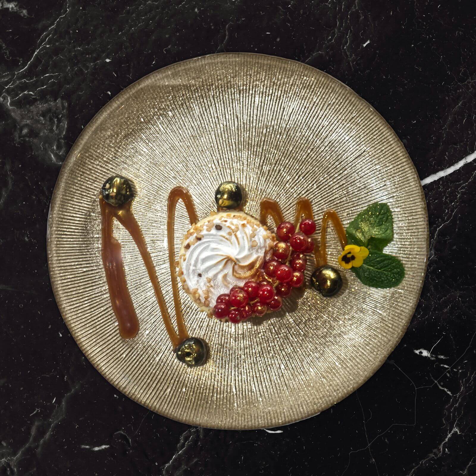 Dessert met meringue, rode bessen, karamel en munt op een goudkleurig bord.