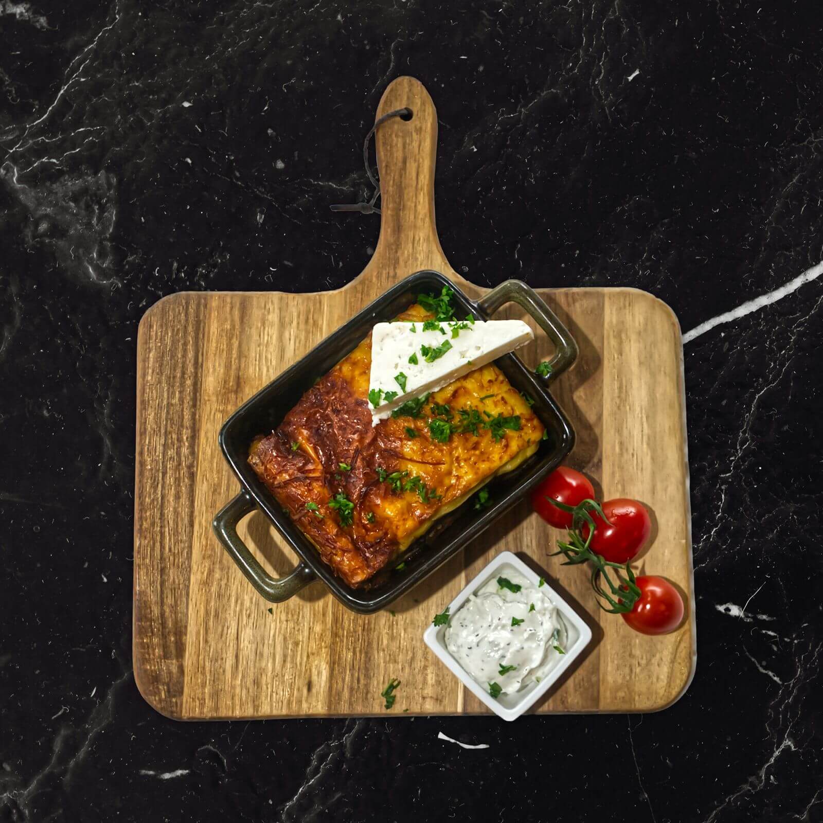 Mousaka in een zwart ovenschaaltje op een houten plank, met een punt feta, tomaatjes en tzatziki.