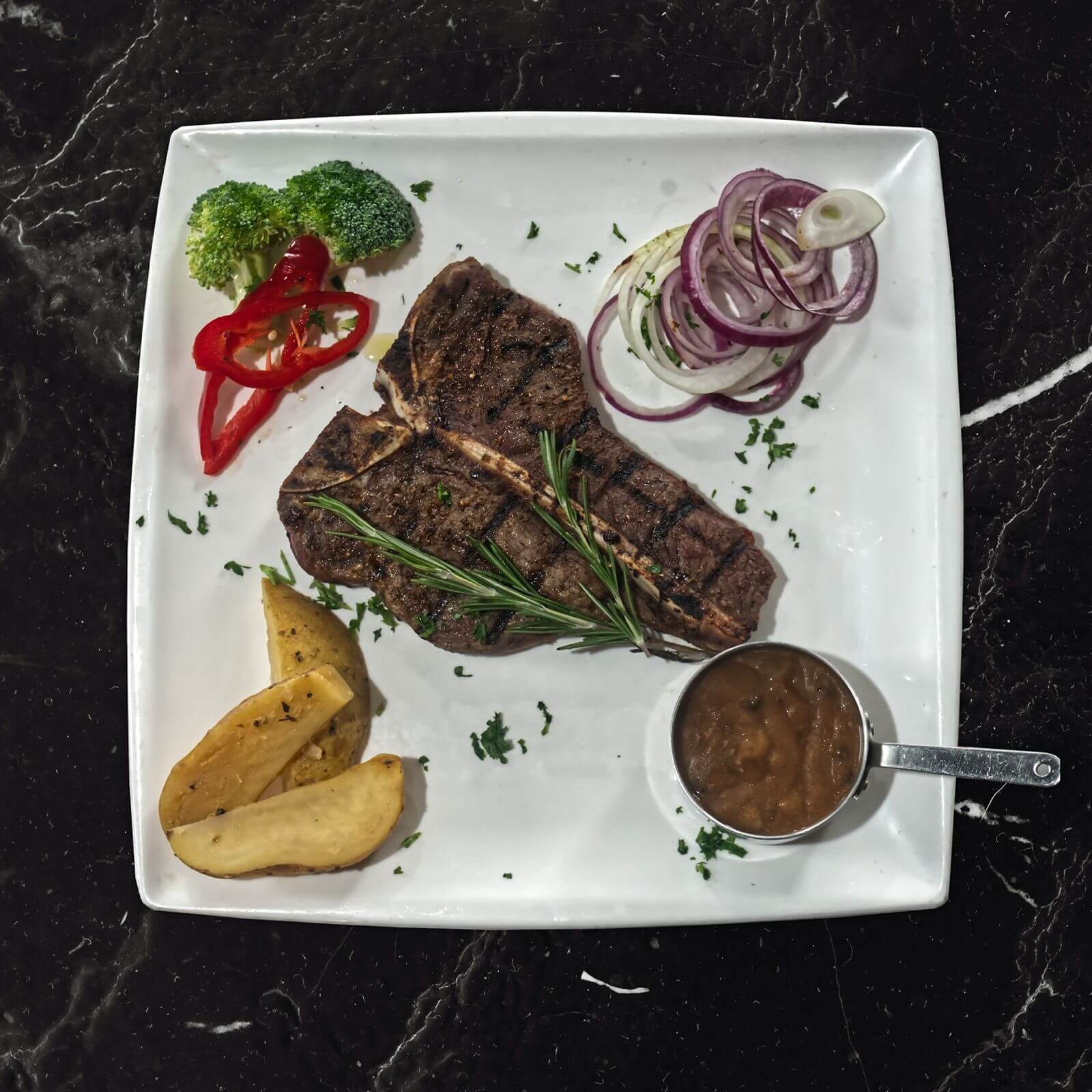 Gegrilde T-Bone steak op een wit vierkant bord met rode ui, broccoli, paprika, aardappelpartjes en een pannetje saus.