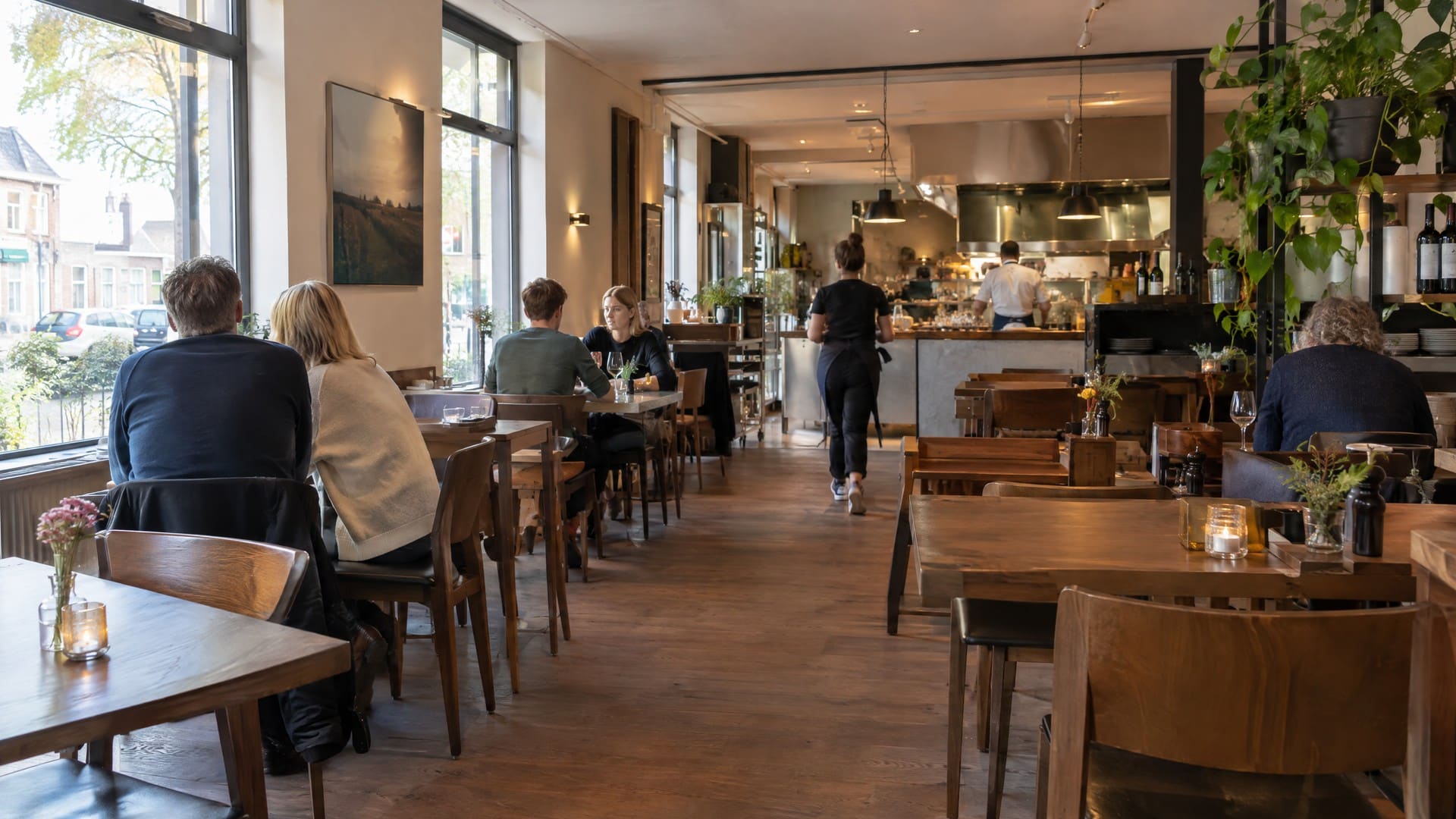 Een warm verlichte restaurantzaal met houten tafels, gasten aan tafel en een open keuken achterin.